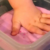A child's hand in a container of pink flubber.